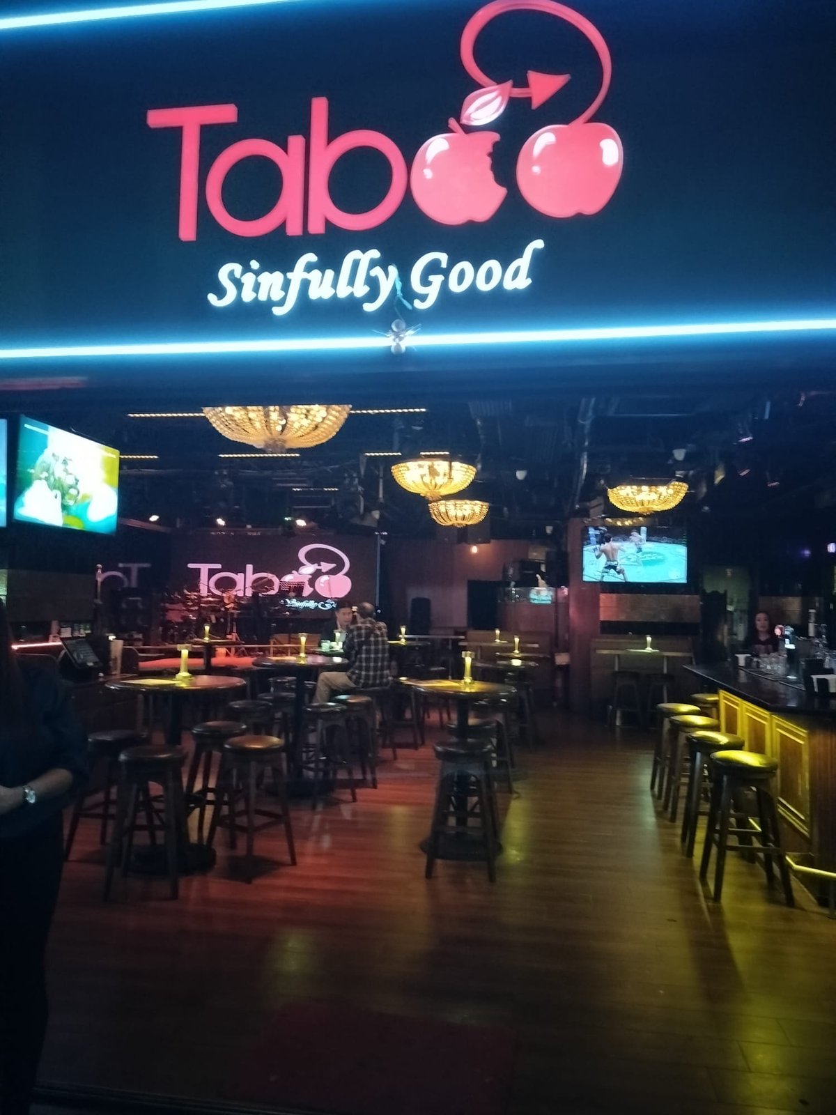 Taboo nightclub interior with pink neon sign, golden ceiling lights, bar seating, and screens displaying neon logo