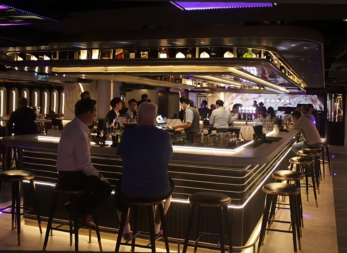 Modern bar interior with illuminated counter, seated patrons, and staff serving drinks in an upscale nightclub setting